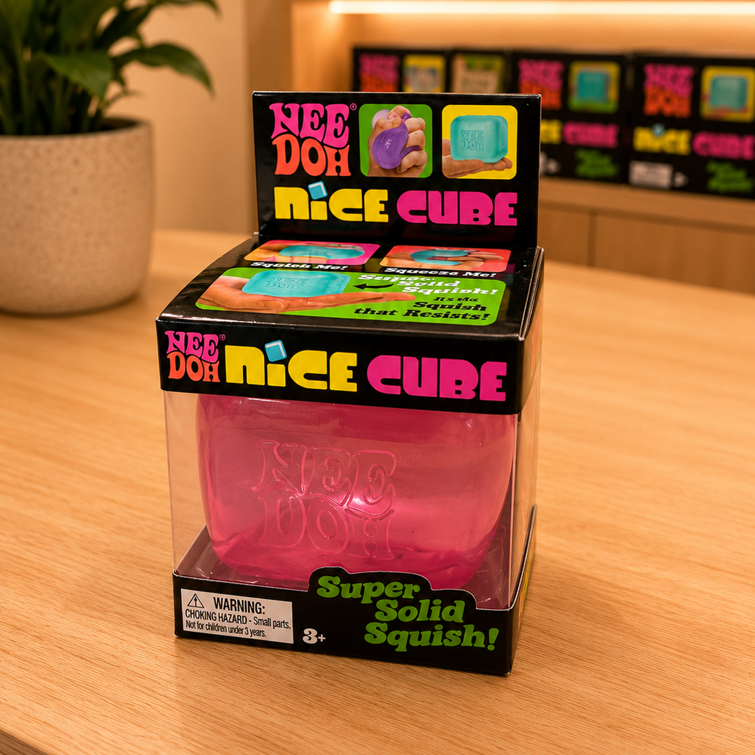 Pink 'Nee Doh Nice Cube' packaging on a wooden surface with a store shelf in the background.